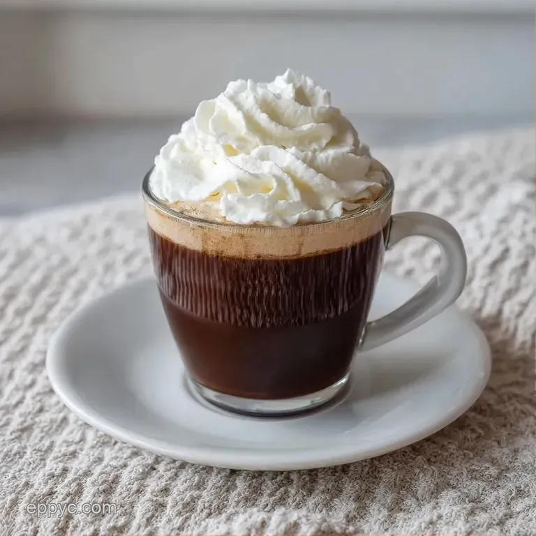A clear glass mug of dark coffee topped with thick cream, set on a rustic wooden coaster with a cinnamon stick.